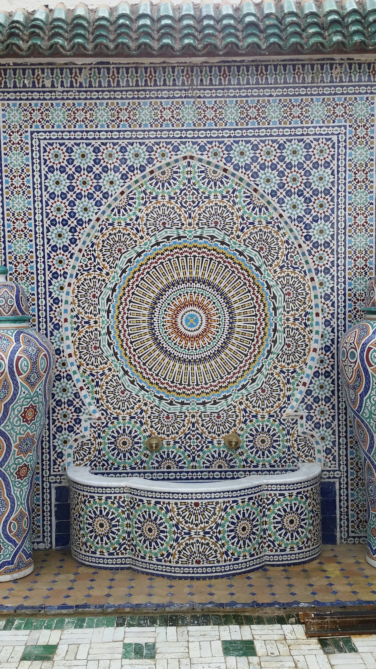 Ornate zellige-tiled Moroccan fountain wall with brass taps, traditional mosaic pattern typical of hammam and riad spa interiors